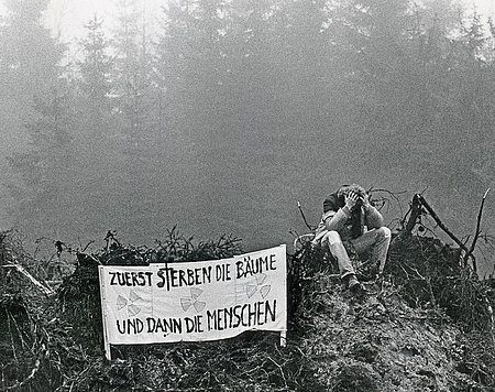 Schwarzweißfoto: im Hintergrund Waldbäume, im Vordergrund ein Haufen umgestürzter Baumwurzeln, darauf ein Mann sitzend mit Plakat: Zuerst sterben die Bäume und dann die Menschen.
