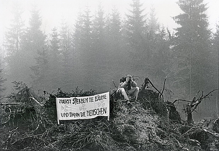 Schwarzweißfoto: im Hintergrund Waldbäume, im Vordergrund ein Haufen umgestürzter Baumwurzeln, darauf ein Mann sitzend mit Plakat: Zuerst sterben die Bäume und dann die Menschen.